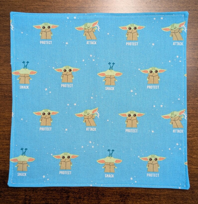 May include: A blue fabric square with a repeating pattern of a green character with different expressions. The character is surrounded by white stars. The text "PROTECT", "ATTACK", and "SNACK" appears on the fabric.
