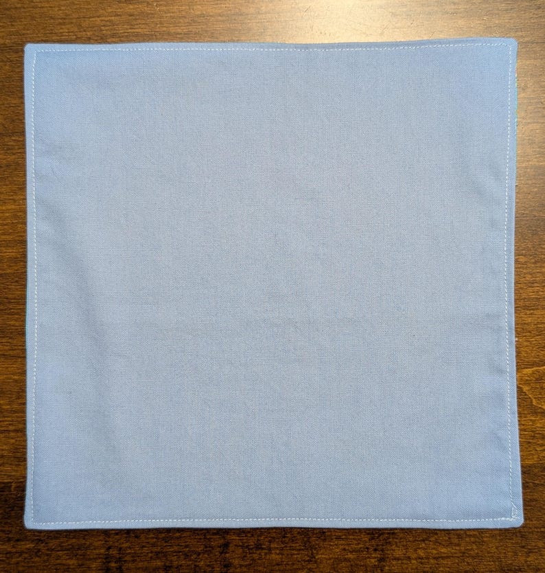 May include: A plain light blue fabric square with white stitching around the edges.
