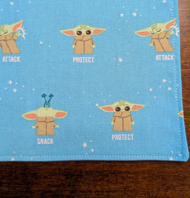 May include: Blue fabric with a repeating pattern of a green alien character with different expressions. The alien is wearing a brown robe and has large ears. The text "Attack", "Protect", and "Snack" appears below the alien characters.