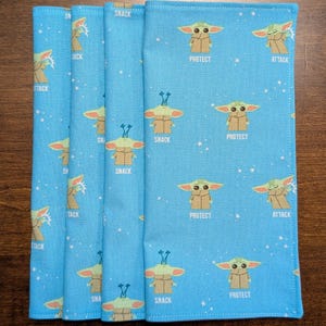 May include: Four blue fabric napkins with a repeating pattern of a green alien character with different expressions. The alien character is surrounded by white stars. The text "Snack", "Protect", and "Attack" appears on the napkins.