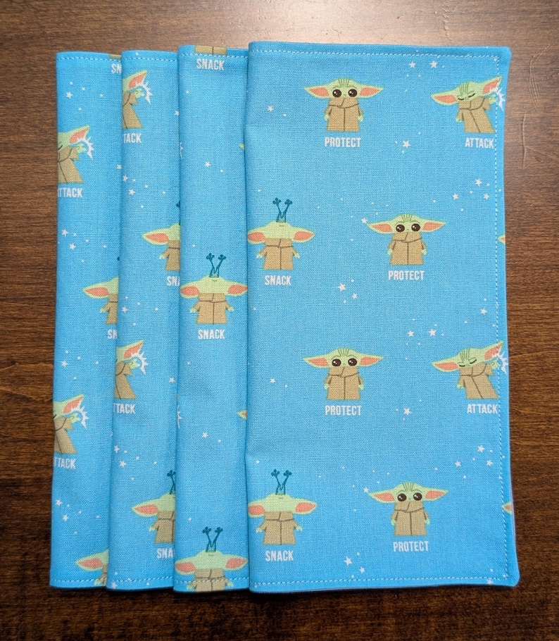 May include: Four blue fabric napkins with a repeating pattern of a green alien character with different expressions. The alien character is surrounded by white stars. The text "Snack", "Protect", and "Attack" appears on the napkins.