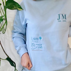 Puede incluir: Sudadera azul claro con un diseño bordado en blanco que presenta una silla, nubes, un globo y el texto "You should be dancing with your heart". La sudadera también tiene el texto "JM est. 1995" bordado en el lado derecho del pecho.