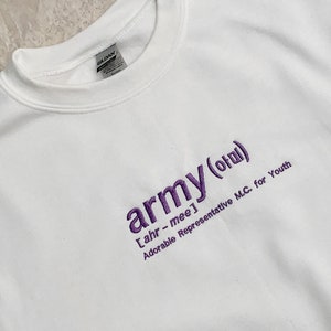 Puede incluir: Sudadera blanca de cuello redondo con la palabra "army" bordada en morado, junto con caracteres coreanos y la frase "Adorable Representative M.C. for Youth". Estilo casual y cómodo.