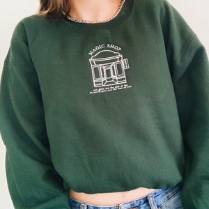 Puede incluir: Una sudadera verde oscuro recortada con un diseño bordado en blanco de una tienda con el texto "MAGIC SHOP" encima. El texto debajo de la tienda dice "you give me the best of you, so I'll give you the best of me."