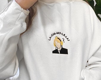 Könnte beinhalten: Weißer Pullover mit einem gestickten Design in Gelb und Schwarz einer Person mit Kopfhörern und dem Text "LA-CHI-MO-LA-LA?"