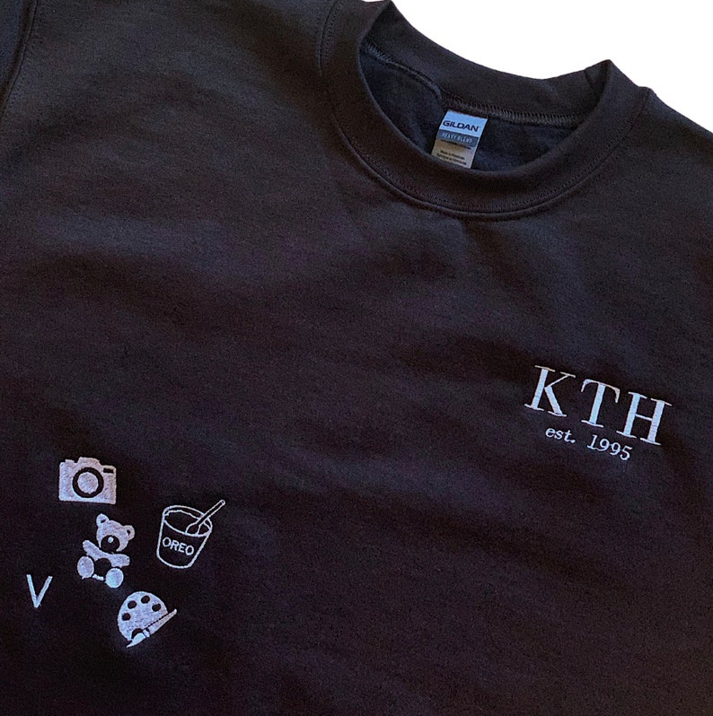 Puede incluir: Sudadera negra con texto blanco que dice "KTH est. 1995". La sudadera tiene un peque&ntilde;o gr&aacute;fico blanco de una c&aacute;mara, un osito de peluche, una taza con una cuchara y una paleta de pintura en la parte delantera.