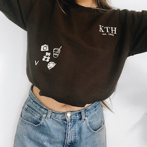 Puede incluir: Una sudadera marr&oacute;n recortada con el texto "KTH est. 1995" en la parte delantera. La sudadera tiene un peque&ntilde;o gr&aacute;fico de una c&aacute;mara, un osito de peluche, una taza con una pajita y una paleta de pintura.