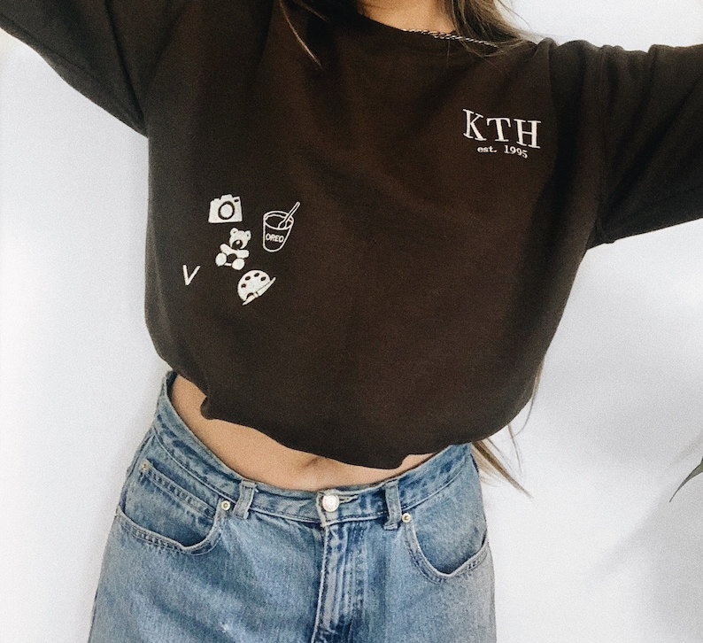 Puede incluir: Una sudadera marr&oacute;n recortada con el texto "KTH est. 1995" en la parte delantera. La sudadera tiene un peque&ntilde;o gr&aacute;fico de una c&aacute;mara, un osito de peluche, una taza con una pajita y una paleta de pintura.