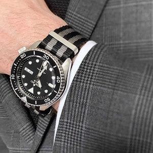 以下が含まれることがあります： 黒とグレーのストライプのナイロン製腕時計バンドにシルバーのバックルが付いています。バンドは、黒と銀の腕時計を着けている手首に巻かれています。腕時計は、黒い文字盤に白い目盛りが付いています。