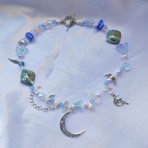 Könnte beinhalten: Eine zarte silberne Halskette mit einem Halbmond-Anhänger, bestehend aus verschiedenen blauen, rosa und weißen Perlen, darunter Schmetterlings- und Engelsflügel-Anhänger.