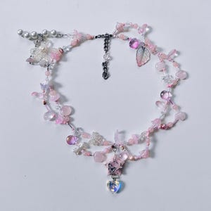 May include: A multi-strand beaded necklace featuring pink and clear glass beads, small white pearls, and silver-toned charms including butterflies, a star, and a heart-shaped pendant. The necklace includes a silver-toned metal chain extender.