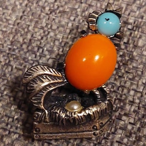 May include: A silver brooch in the shape of a rooster with an orange and turquoise stone. The rooster is standing on a base with a pearl.