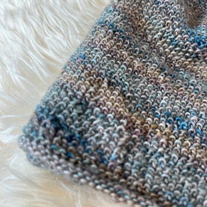 May include: A hand-knitted beanie crafted from variegated yarn featuring a blend of earthy brown, soft grey, and vibrant blue tones. The fabric displays a consistent garter stitch pattern throughout, creating a textured surface with a multi-colored appearance.