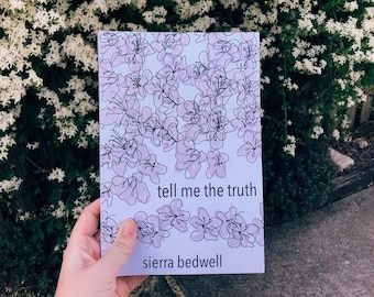 May include: A book cover with a floral design in shades of pink and black, with the title "tell me the truth" and the author's name, sierra bedwell. The book is held in front of a backdrop of white flowers.