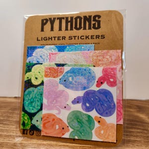 May include: A pack of holographic vinyl lighter stickers featuring colorful snake illustrations. The stickers are in various colors, including blue, pink, green, and orange, with glitter accents. The packaging reads "PYTHONS" and "LIGHTER STICKERS".