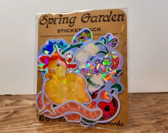 May include: A sticker pack titled "Spring Garden" featuring several holographic stickers. The stickers include illustrations of a duck, flowers, and a ladybug. The packaging is brown with the words "Spring Garden" in a script font.
