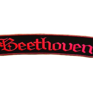 Puede incluir: Un parche bordado rectangular con esquinas redondeadas que presenta un fondo negro y un borde rojo. La palabra Beethoven está cosida con hilo rojo en el centro en una fuente de estilo gótico, creando un diseño audaz y de alto contraste.