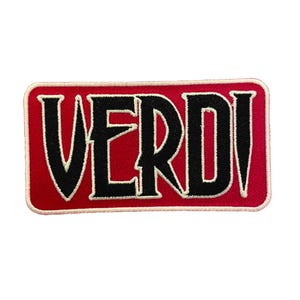 May include: A rectangular embroidered patch featuring the word VERDI in bold black lettering with a white outline. The text is set against a vibrant red background, and the entire patch is finished with a clean, light-coloured stitched border.