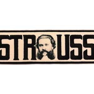 Pode incluir: Um emblema retangular bordado com fundo creme e borda preta. A palavra STRAUSS está escrita em letras pretas em negrito, com a letra A substituída por um retrato detalhado em preto e branco de um homem com bigode.