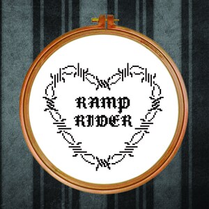 May include: A white embroidery hoop with black thread depicting a barbed wire heart with the text "RAMP RIDER" inside.