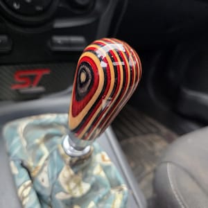 May include: A gear shift knob featuring a polished, multi-layered wood design with alternating bands of red, black, and natural tan colors. The knob is mounted on a silver metal base, positioned above a light blue fabric boot with a white floral pattern.