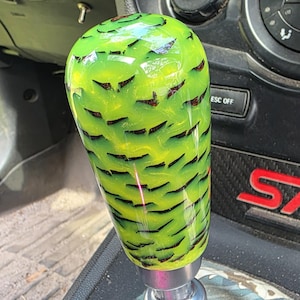 Puede incluir: Un pomo de cambio verde y negro personalizado con un patrón único. El pomo es cilíndrico con una parte superior redondeada y está montado sobre una base de metal plateado. El fondo muestra el interior de un coche.