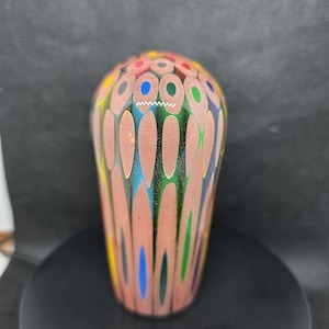 Könnte beinhalten: Ein dekoratives, abgerundetes Objekt, das aus zahlreichen farbigen Bleistiften gefertigt wurde. Die Oberseite zeigt ein Mosaik aus Bleistiftspitzen in verschiedenen Farben, während die Seiten die Holzmaserung zeigen. Das Objekt befindet sich vor einem dunklen Hintergrund.