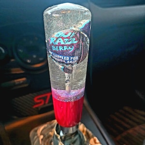 Könnte beinhalten: Ein klarer, zylindrischer Schalthebelknauf mit roter Basis und einer Süßigkeiten-Grafik im Inneren. Die Grafik zeigt "Razz Berry" in Blau und Pink. Der Knauf ist in einem Auto eingebaut.