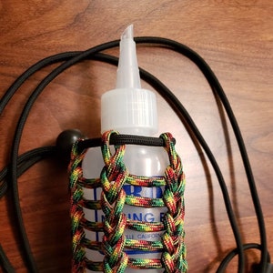May include: A clear bottle with a white nozzle is encased in a colorful braided paracord holder. The holder features red, yellow, green, and black strands. A black cord with a clasp is attached, suggesting it can be worn.