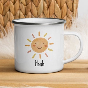 K&ouml;nnte beinhalten: Wei&szlig;e Email Tasse mit einer l&auml;chelnden Sonnen-Illustration und dem Namen "Noah" darunter. Die Tasse hat einen silbernen Rand und einen schwarzen Griff.
