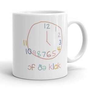 May include: White ceramic mug with a hand-drawn clock design. The clock face has numbers 1-12 and the words "of öä klak" written below.