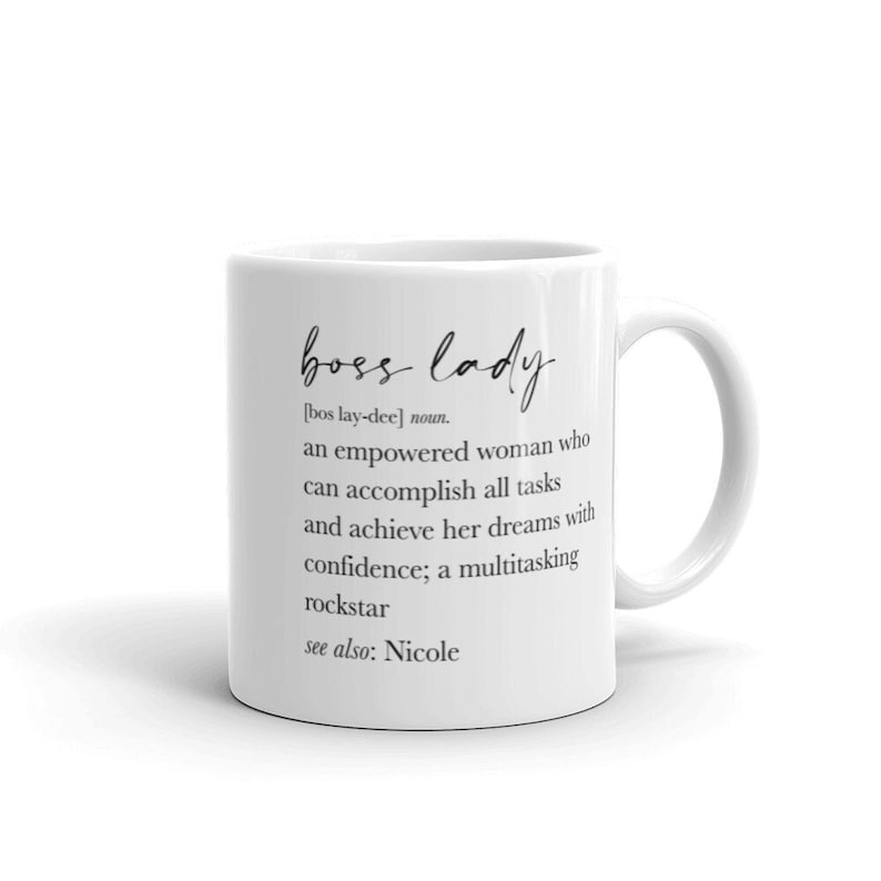 May include: White ceramic mug featuring the words "boss lady" in a script font. The mug includes a definition of "boss lady" and the text "see also: Nicole". The mug has a curved handle.