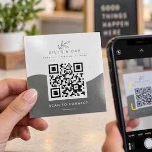 May include: A square white card featuring the black text River & Oak, Home Lifestyle More, and Scan to Connect. A black QR code is centered on the card, which has a gray wavy graphic element at the bottom. A smartphone is positioned to scan the card.