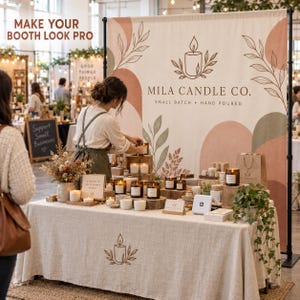 May include: A large fabric trade show backdrop featuring the text "MILA CANDLE CO. SMALL BATCH HAND POURED" with a candle logo and botanical illustrations in muted earth tones, displayed behind a table covered in a linen cloth with candles.