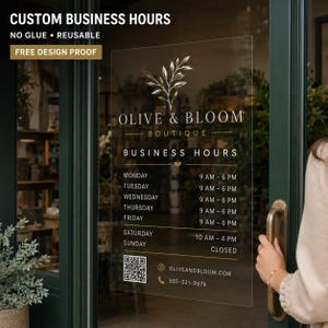 May include: A clear, reusable business hours decal applied to a glass door. The sign features gold text and a botanical logo for Olive & Bloom Boutique, listing operating hours from Monday to Sunday, a QR code, a website address, and a phone number.