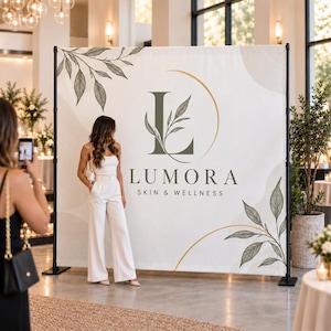 May include: A large rectangular step and repeat banner featuring a white background with olive green botanical leaf illustrations, gold curved lines, and the text Lumora Skin & Wellness centered with a large letter L integrated with a leaf design.