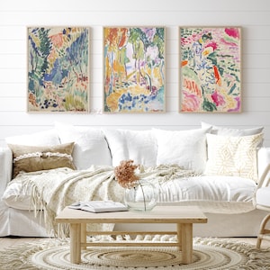 May include: Three abstract paintings in a living room setting. The paintings are framed in wood and feature colourful brushstrokes and abstract shapes. The paintings are hung on a white wall above a white sofa with cushions and a throw blanket. A wooden coffee table sits in front of the sofa.