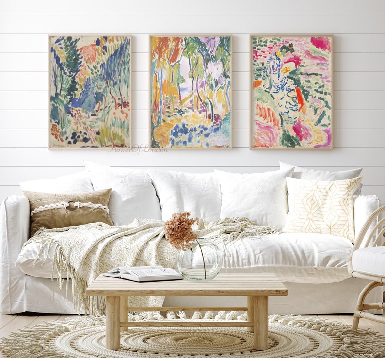May include: Three abstract paintings in a living room setting. The paintings are framed in wood and feature colourful brushstrokes and abstract shapes. The paintings are hung on a white wall above a white sofa with cushions and a throw blanket. A wooden coffee table sits in front of the sofa.