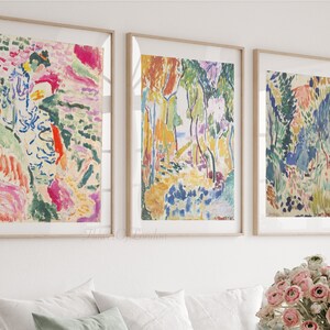 May include: Three framed abstract paintings with colourful brushstrokes and a landscape theme. The paintings are in a white room with a bouquet of pink flowers in the foreground.