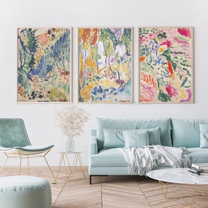 May include: Three abstract landscape paintings in a living room setting. The paintings are framed and feature a colourful, impressionistic style with brushstrokes of blue, green, yellow, pink, and red. The paintings are hung on a white wall above a light blue sofa with white throw pillows and a white throw blanket.