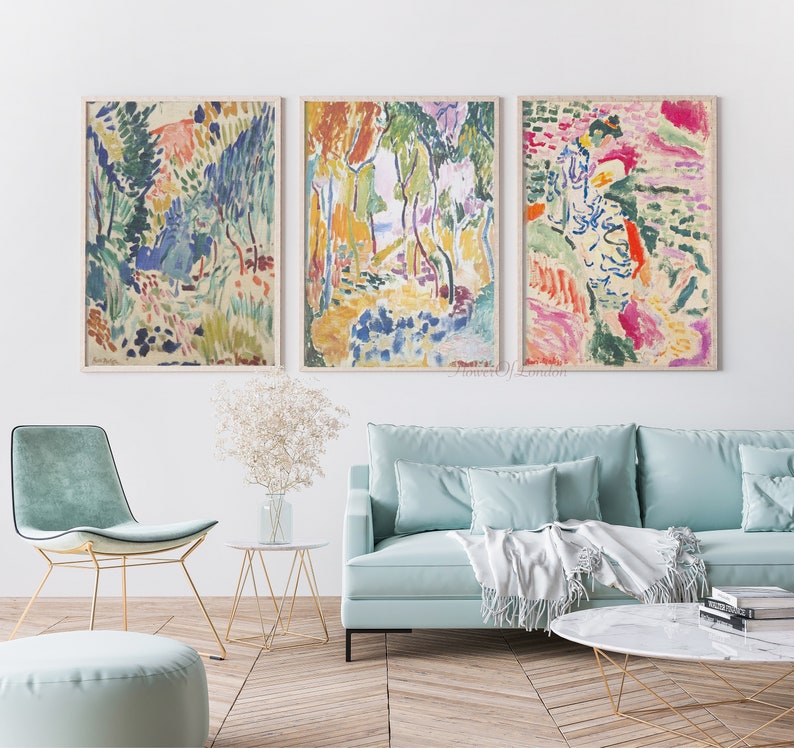 May include: Three abstract landscape paintings in a living room setting. The paintings are framed and feature a colourful, impressionistic style with brushstrokes of blue, green, yellow, pink, and red. The paintings are hung on a white wall above a light blue sofa with white throw pillows and a white throw blanket.