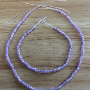 May include: Two strands of beaded jewelry in a soft lavender hue. The beads are small and strung on a clear, thin cord. One strand is a necklace, and the other is a bracelet. The jewelry is displayed on a light brown wooden surface.