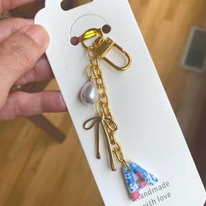 May include: Gold-toned keychain with a chain, clasp, and charms. Charms include a faux pearl, a bow, and a letter "A" in blue and pink patterned fabric. The card reads "handmade with love" with a red heart.
