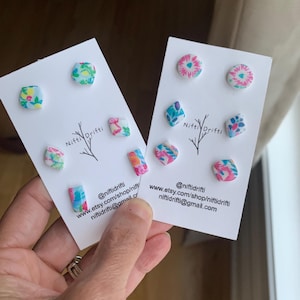 May include: Two white earring display cards with colorful floral stud earrings. The cards have the text "Nifti Drifti" and social media contact information. The earrings feature pink, blue, and green designs.