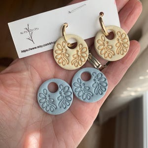 May include: A pair of cream-colored earrings with gold hoops and a floral design, along with two light blue earring pendants. The earrings are displayed on a white card with the text "Niffi Drifti" and a website address.