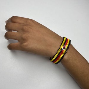 May include: A beaded bracelet with black, yellow, and red stripes and a white circle in the center.