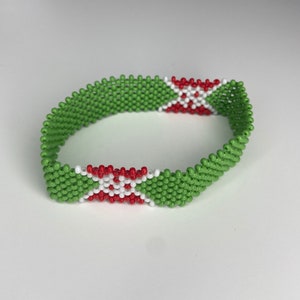 May include: A green beaded bracelet with red and white beads forming a cross pattern. The bracelet is made of small, round beads.