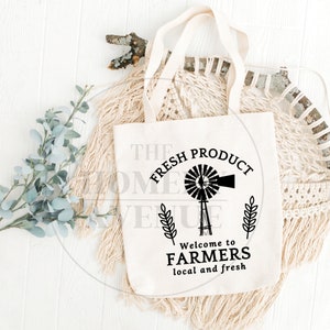 Pu&ograve; includere: Una borsa di tela bianca con testo nero e un grafico di un mulino a vento. Il testo dice "The Fresh Product" con uno stelo di grano su entrambi i lati del mulino a vento. Sotto il mulino a vento, il testo dice "Welcome to Farmers local and fresh".