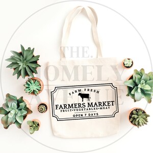 May include: A white canvas tote bag with black text that reads "Farm Fresh Farmers Market Fruit Vegetables Meat Open 7 Days". A black silhouette of a cow is above the text. The bag is surrounded by small potted succulents.