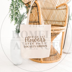 May include: A white canvas tote bag with brown text that reads "Fresh Cut Flowers Open Daily Locally Grown". The bag is hanging on a wicker chair.